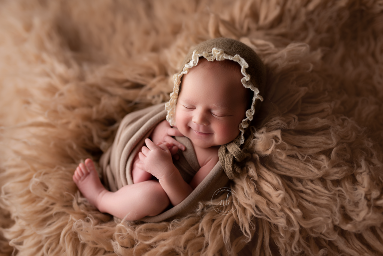 smiling new newborn baby girl in taupe ruffle bonnet on fur rug